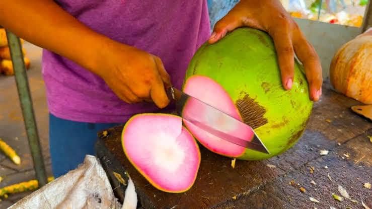Seseorang memotong kelapa muda jenis wulung berwarna hijau dengan bagian dalam berwarna merah muda di atas meja kayu.