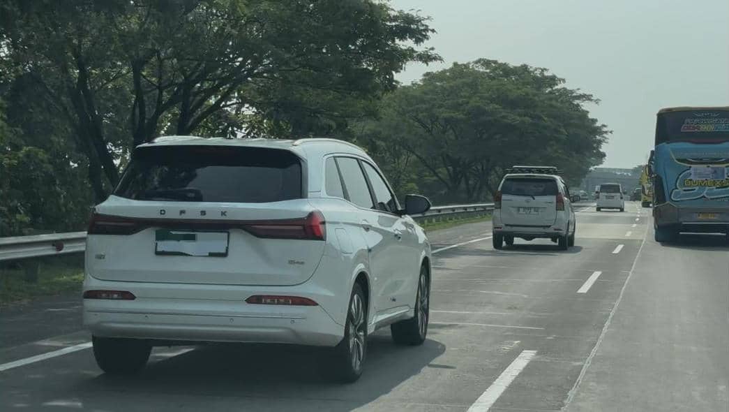Sebuah mobil SUV putih berlogo DFSK melaju di jalan tol bersama beberapa kendaraan lain di siang hari yang cerah.