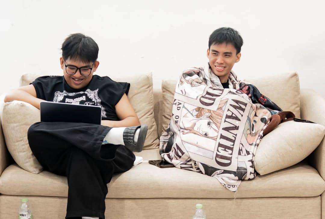 Dua pria duduk santai di sofa, satu menggunakan laptop dan satu lagi tersenyum sambil diselimuti kain bergambar karakter anime.