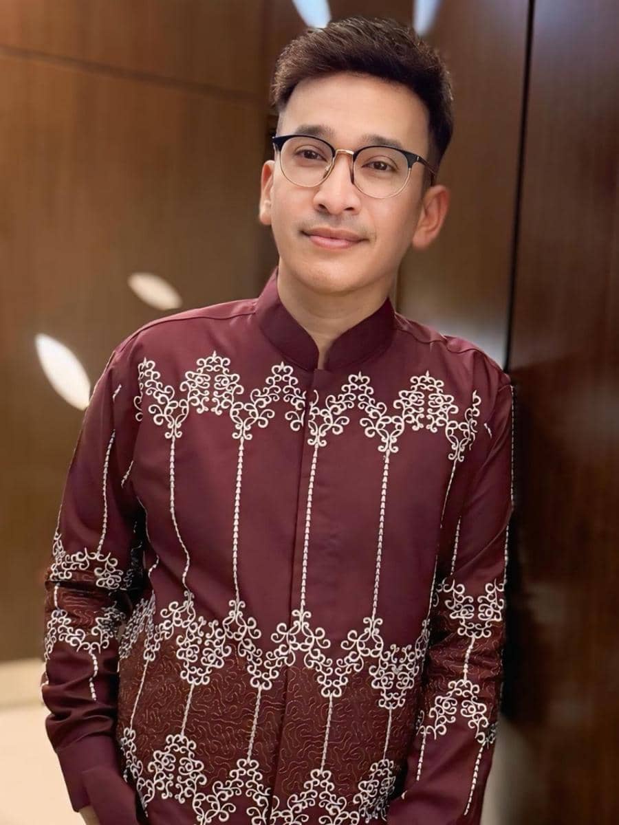 Seorang pria mengenakan kemeja batik berwarna marun dengan motif putih, berdiri di dalam ruangan dengan latar dinding kayu mengilap.