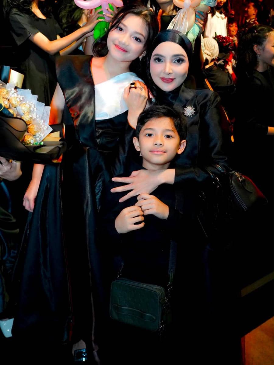 Seorang anak perempuan bersama keluarganya berpose mengenakan busana hitam elegan di acara kelulusan sekolah dasar.