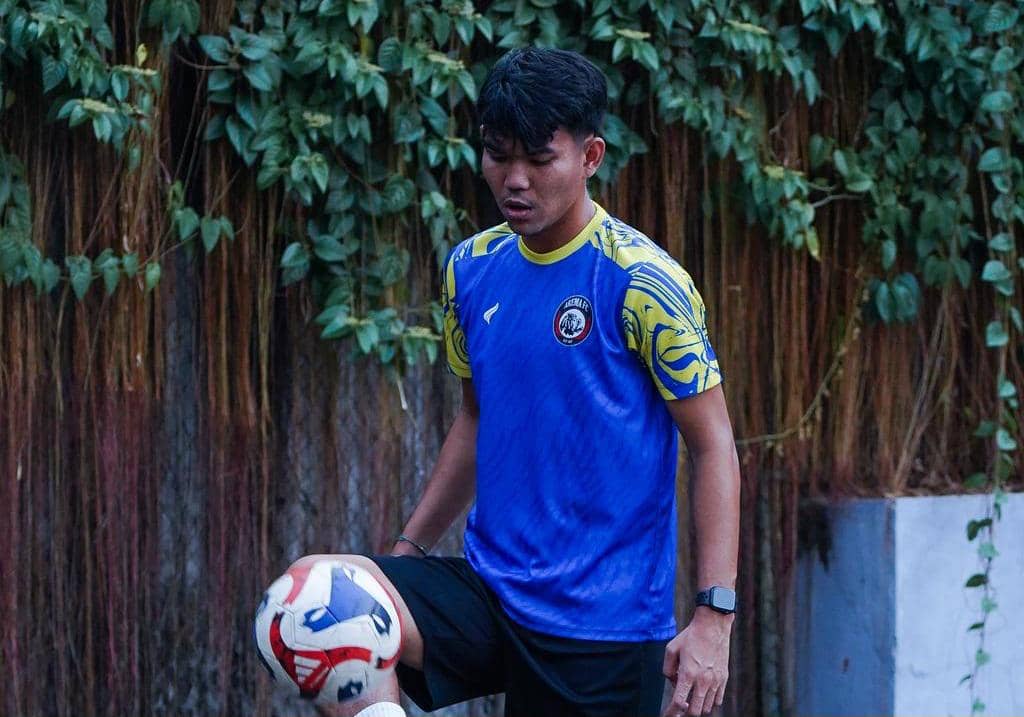 Seorang pemain Arema FC mengenakan jersey biru dengan aksen kuning sedang mengontrol bola di area luar dengan latar tanaman rambat.