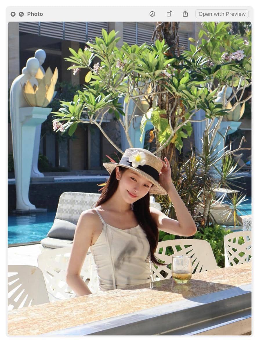 Seorang perempuan mengenakan topi jerami dengan bunga putih duduk di tepi kolam renang di bawah sinar matahari tropis Bali.
