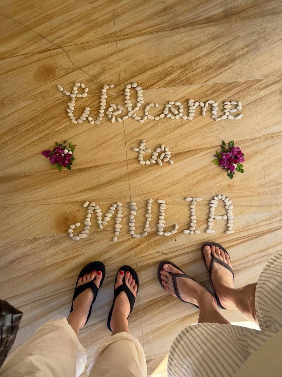 Dua orang mengenakan sandal berdiri di lantai kayu dengan tulisan 'Welcome to Mulia' dari batu kecil dan hiasan bunga ungu.