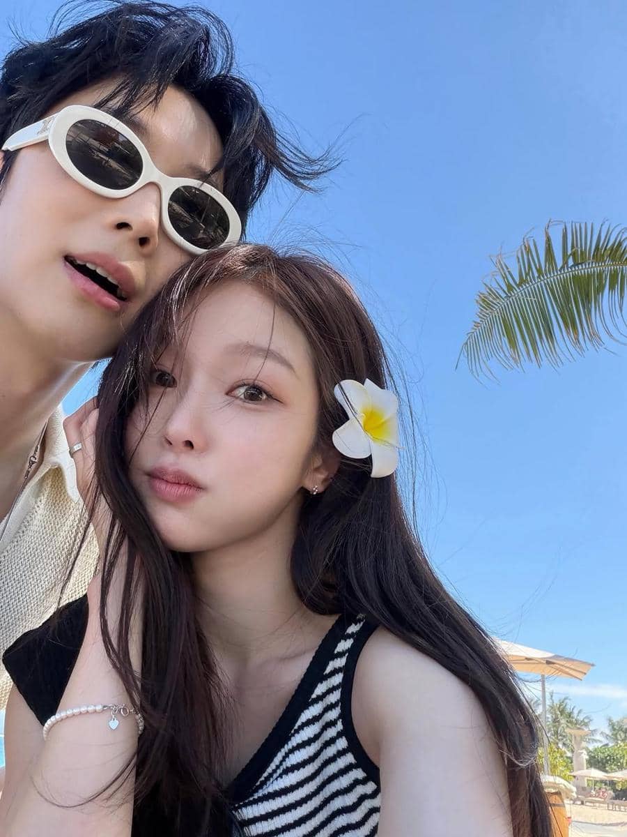 Sepasang anak muda berpose selfie di bawah langit cerah Bali, perempuan mengenakan bunga kamboja di rambutnya dan pria memakai kacamata putih.