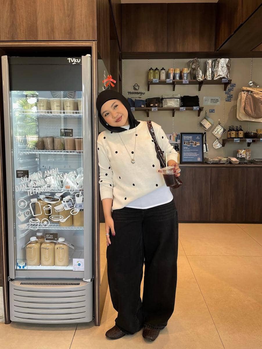 Seorang wanita berhijab mengenakan sweater putih bermotif titik hitam dan celana panjang hitam lebar berpose di depan lemari pendingin kafe.