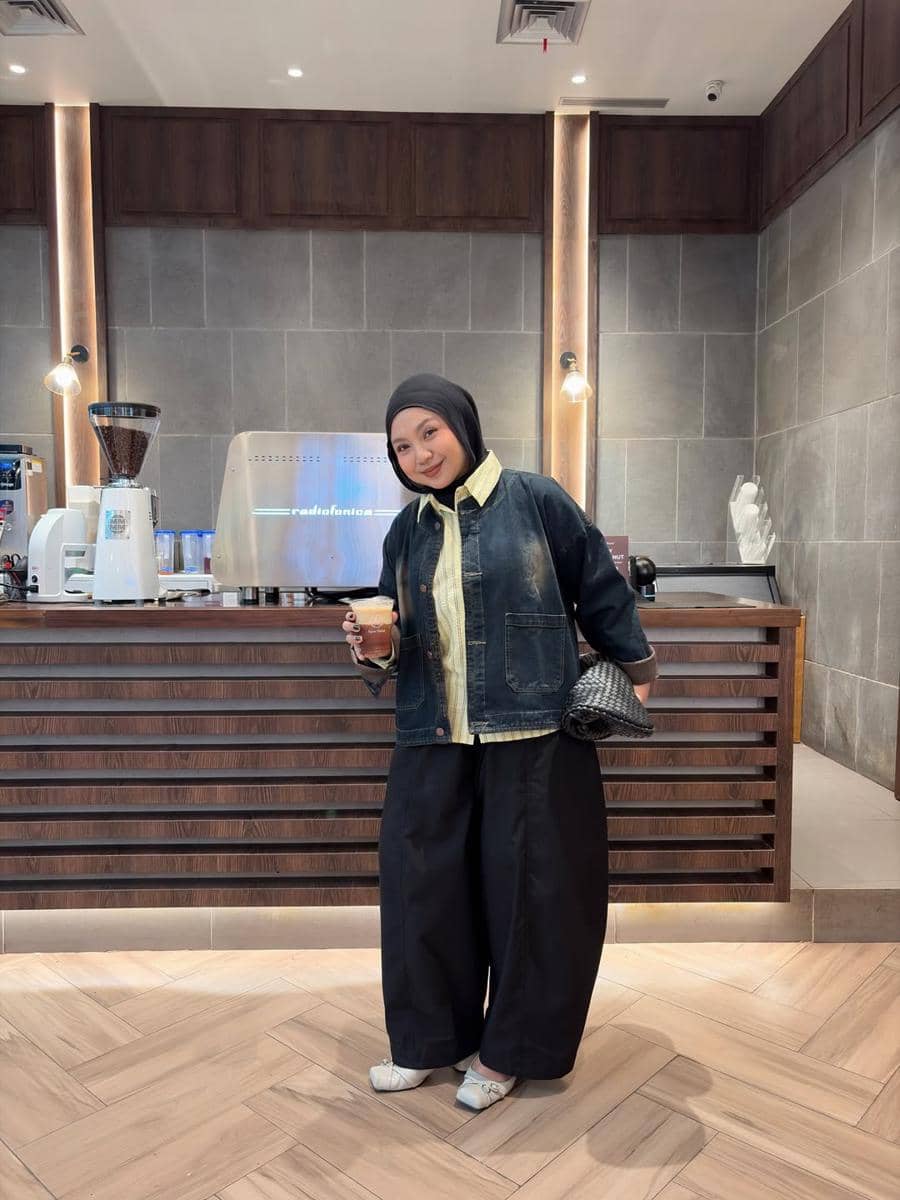 Seorang wanita bergaya kasual mengenakan hijab hitam, jaket denim, dan celana kulot hitam sambil memegang minuman di kafe modern.