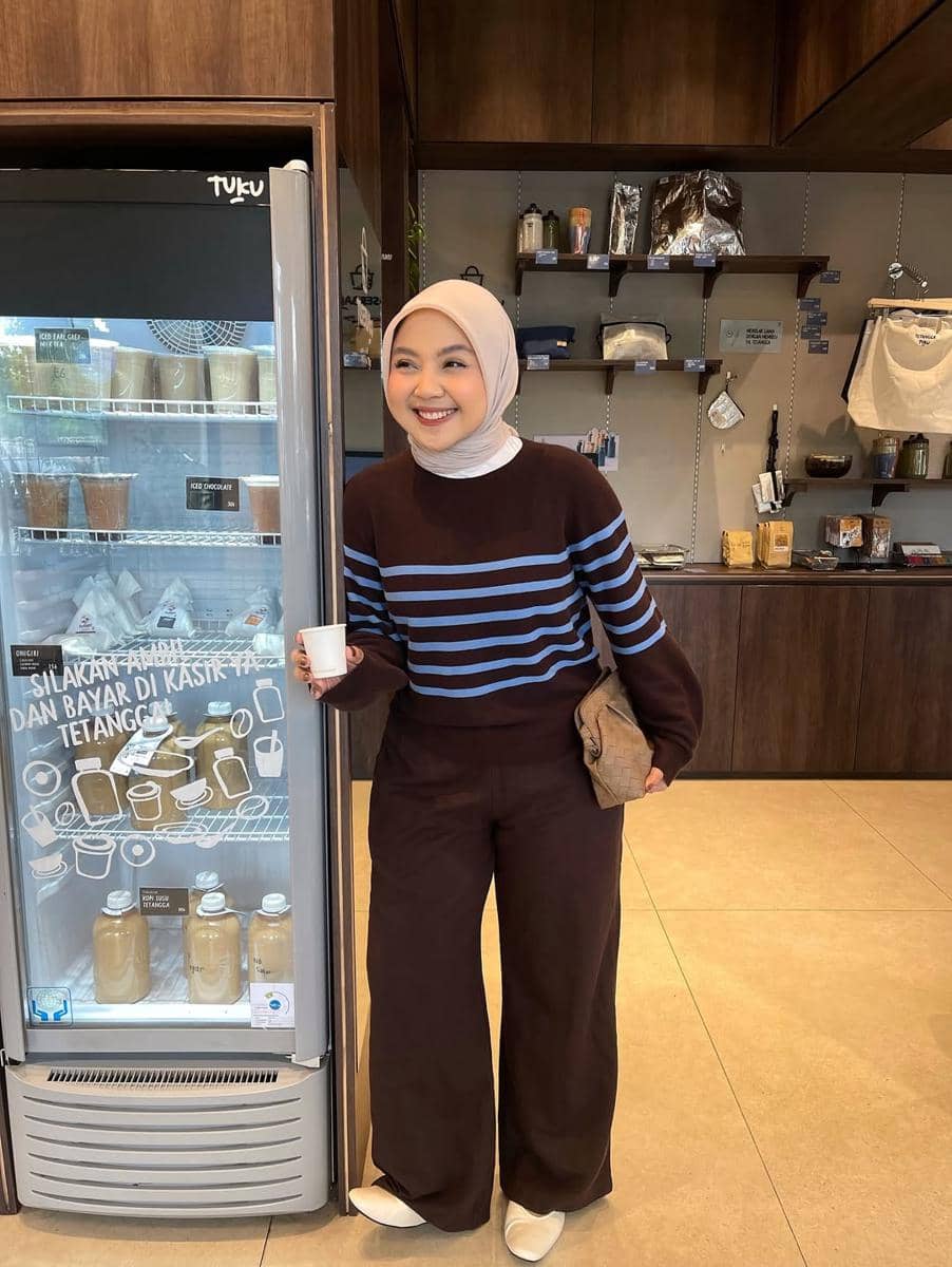 Perempuan berhijab mengenakan sweater cokelat bergaris biru dan celana longgar cokelat sedang berdiri di depan kulkas minuman di kafe.