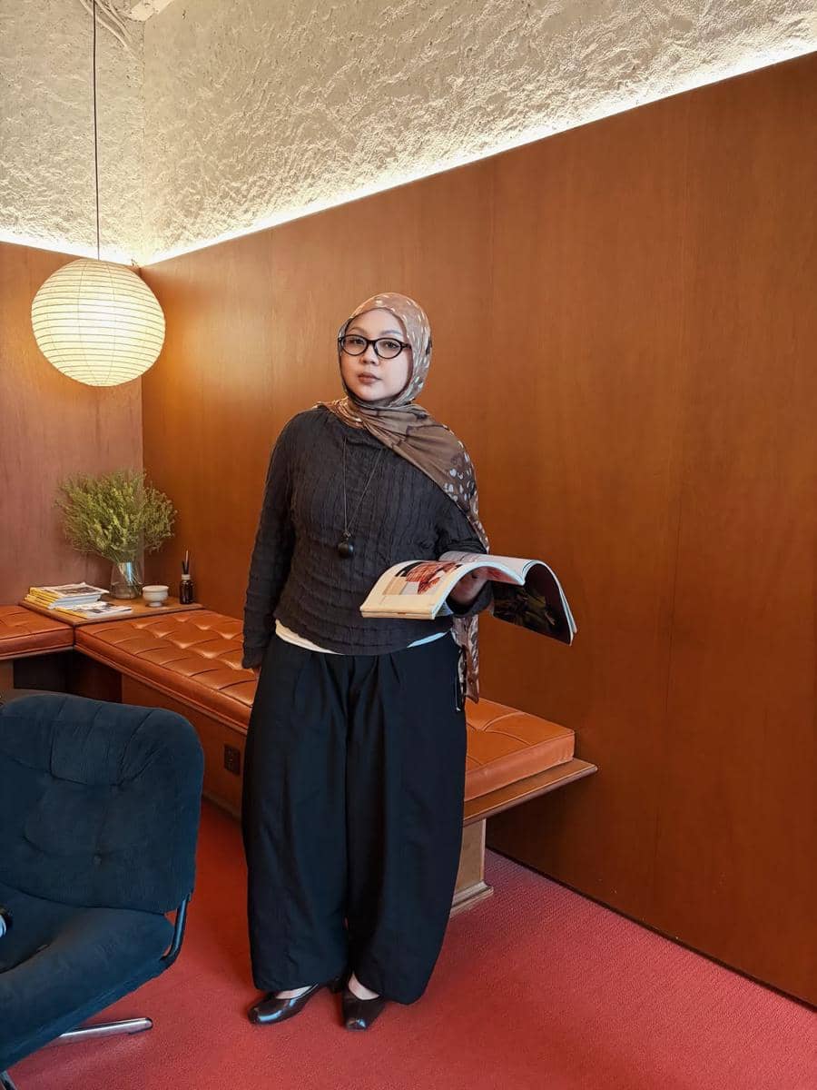 Seorang wanita berhijab mengenakan sweater cokelat tua dan celana hitam longgar sambil memegang majalah di ruangan bergaya modern.