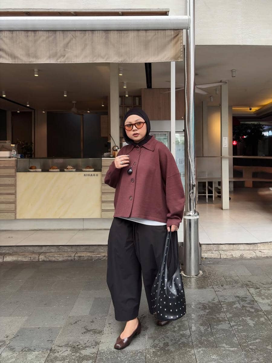 Perempuan berhijab mengenakan jaket marun, celana hitam longgar, dan sepatu cokelat sambil membawa tas hitam di depan kafe.