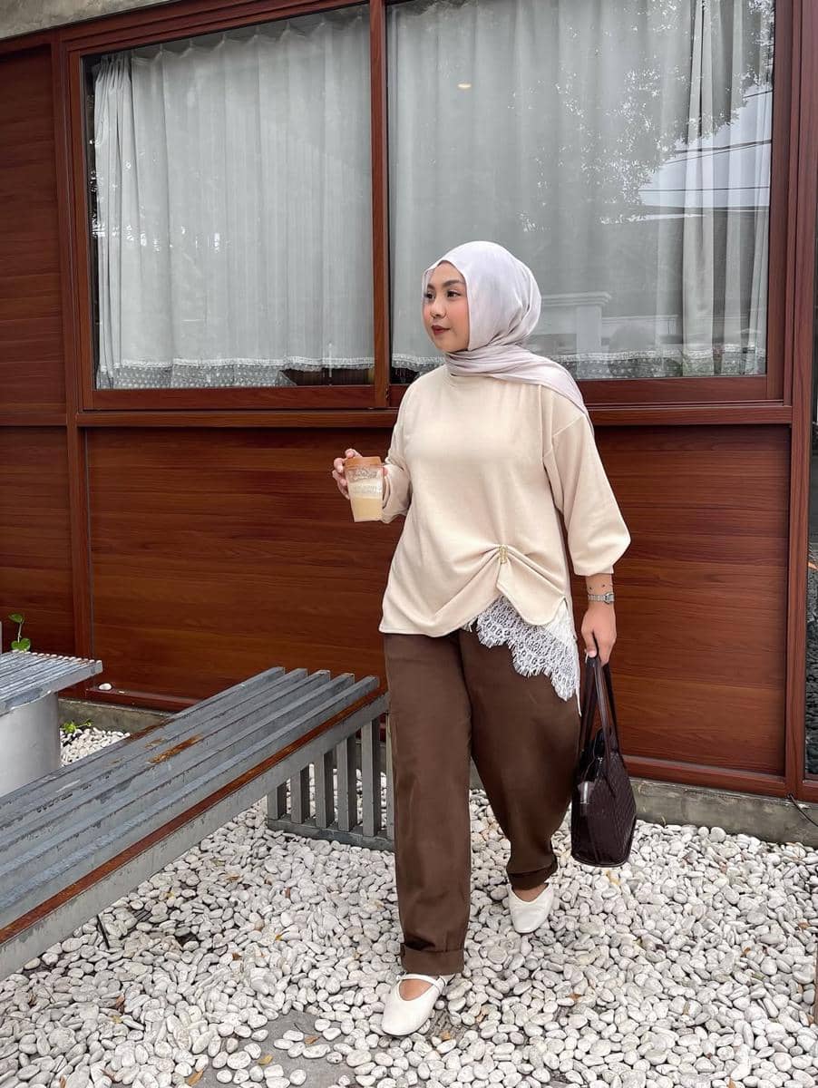 Perempuan berhijab mengenakan atasan krem dan celana cokelat sambil memegang minuman dan tas di area luar dengan dinding kayu.