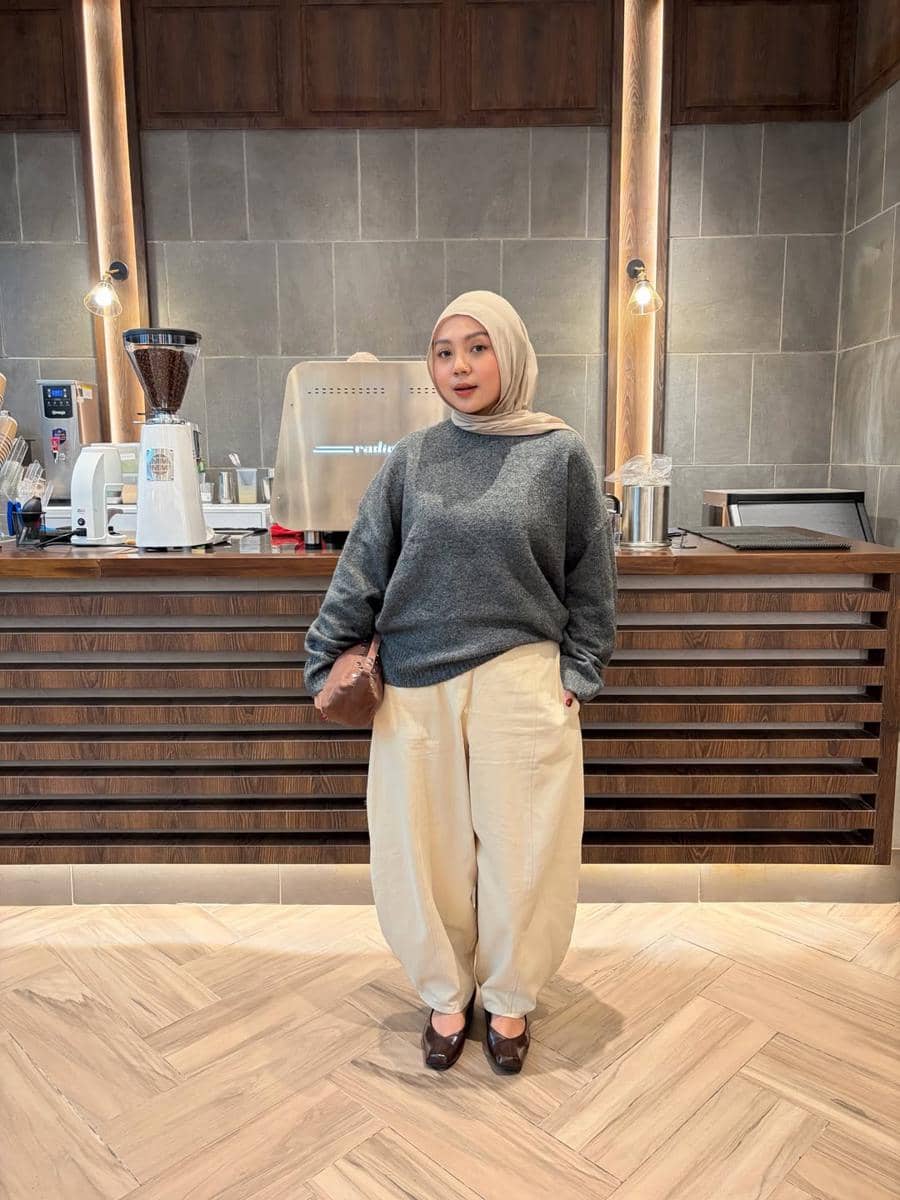 Seorang wanita berhijab mengenakan sweater abu-abu dan celana krem longgar berpose di depan meja bar kafe dengan pencahayaan hangat.