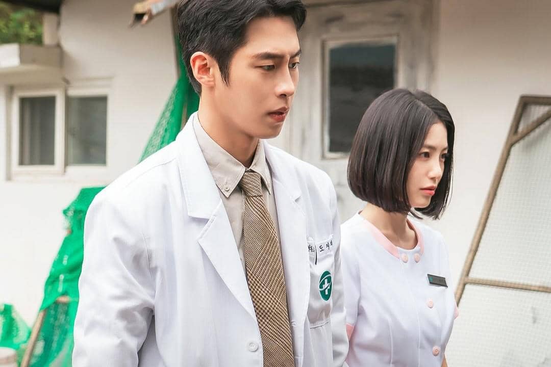 Dua tenaga medis mengenakan jas dan seragam putih berdiri di luar gedung dalam adegan drama Doctor On The Edge.