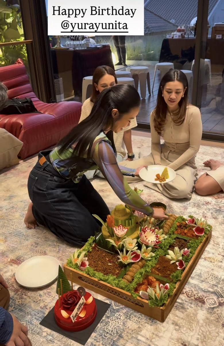 Seorang wanita memotong tumpeng kuning di atas lantai bersama teman-temannya dalam perayaan ulang tahun di dalam ruangan.