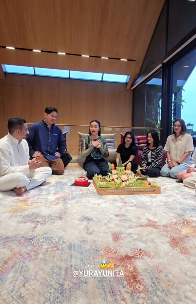 Sejumlah orang duduk melingkar di dalam ruangan modern dengan dekorasi kayu, merayakan ulang tahun dengan tumpeng dan kue di tengah.