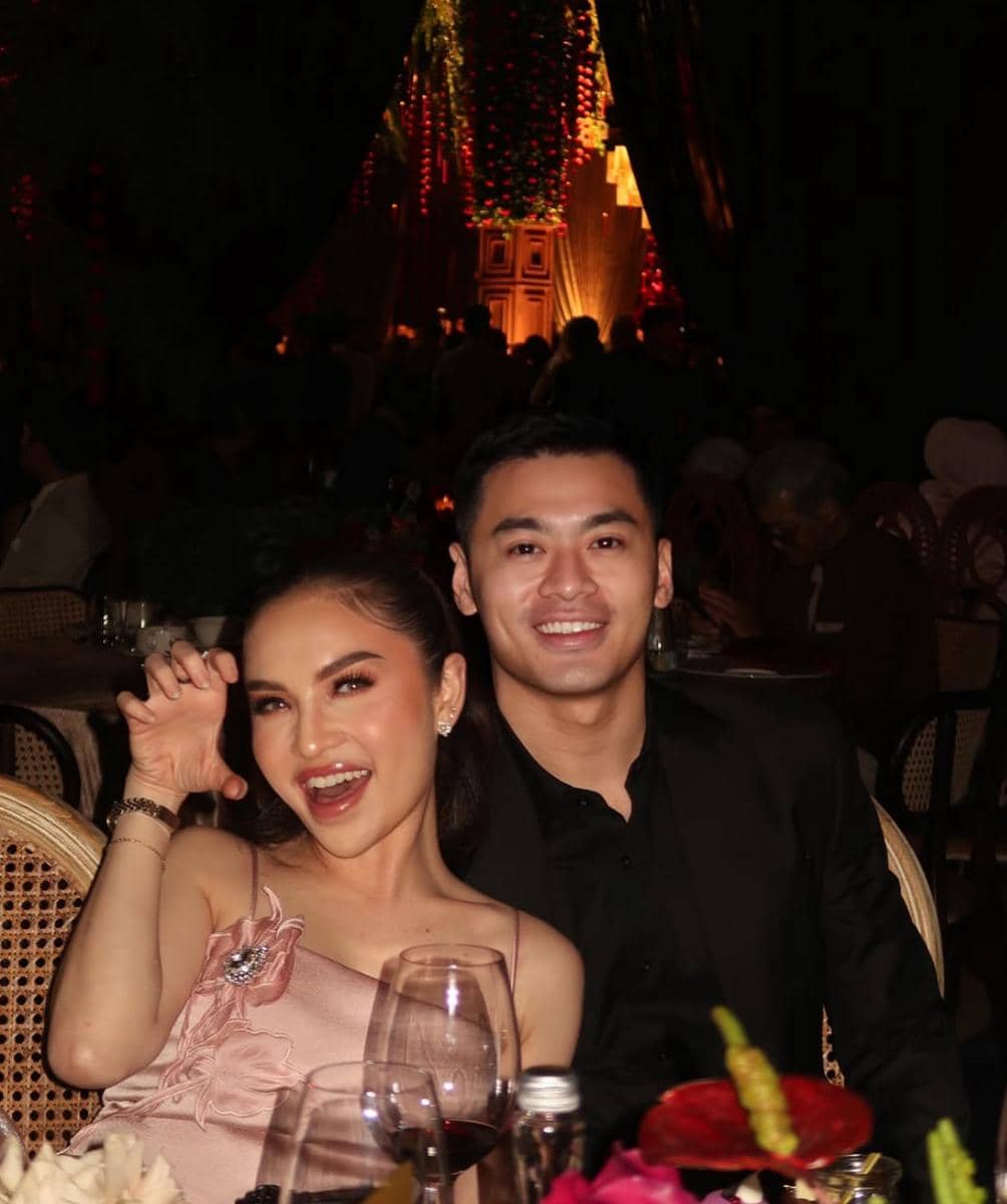 Seorang perempuan bergaun merah muda berpose ceria bersama seorang pria berjas hitam di meja makan dengan suasana pesta malam.