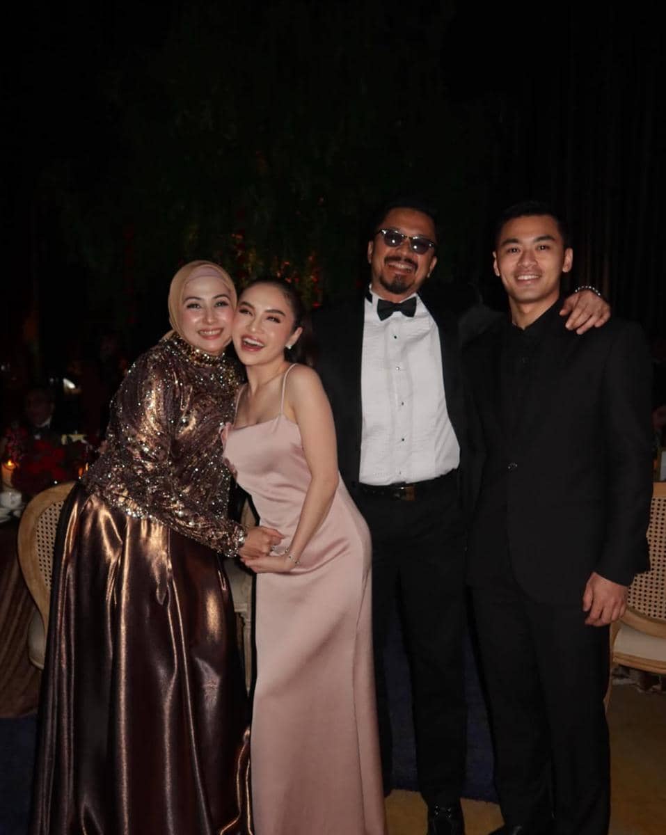 Empat orang berpose bersama di acara malam hari, dua wanita mengenakan gaun elegan dan dua pria mengenakan setelan jas hitam.