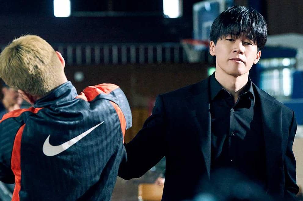 Seorang pria berjas hitam memegang kerah jaket pria lain berambut pirang di dalam ruangan, adegan dari drama Teach You a Lesson.