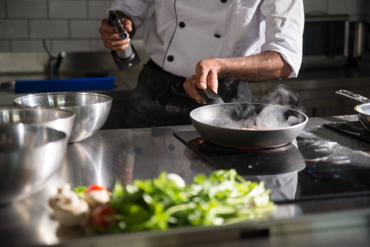 Seorang koki mengenakan seragam putih sedang memasak di dapur profesional dengan wajan di atas kompor dan bahan segar di meja.