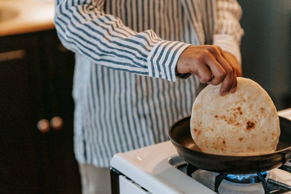 Seseorang mengenakan kemeja bergaris sedang memanggang tortilla di atas teflon di atas kompor gas di dapur.