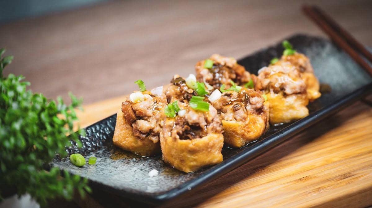 Beberapa potong tahu isi adonan dimsum disajikan di atas piring hitam dengan taburan daun bawang dan saus gurih.