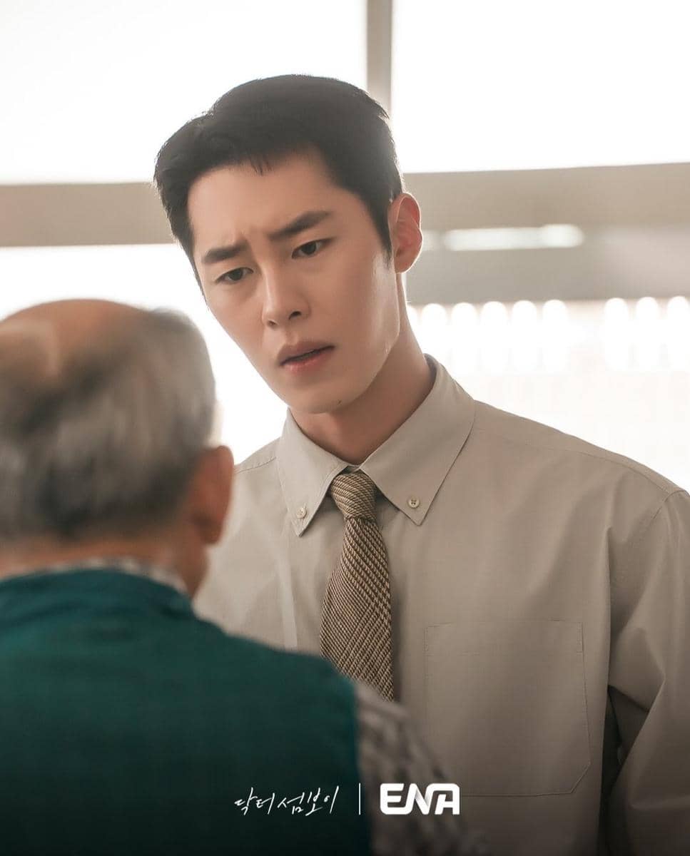 Seorang pria muda mengenakan kemeja dan dasi berbicara serius dengan pria lanjut usia dalam cuplikan drama Doctor on the Edge.