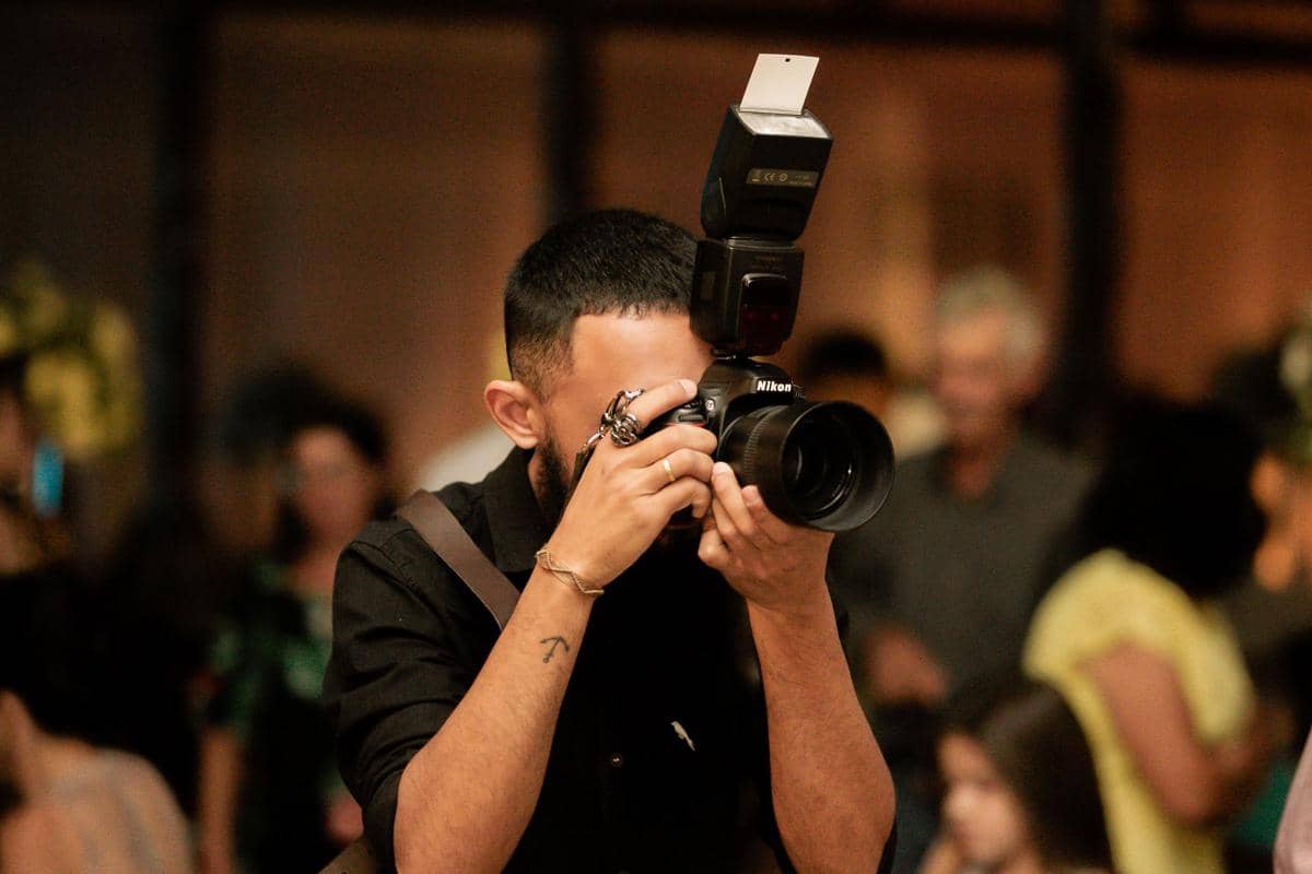 Seorang fotografer memegang kamera profesional dengan lampu kilat eksternal di tengah suasana ramai di dalam ruangan.