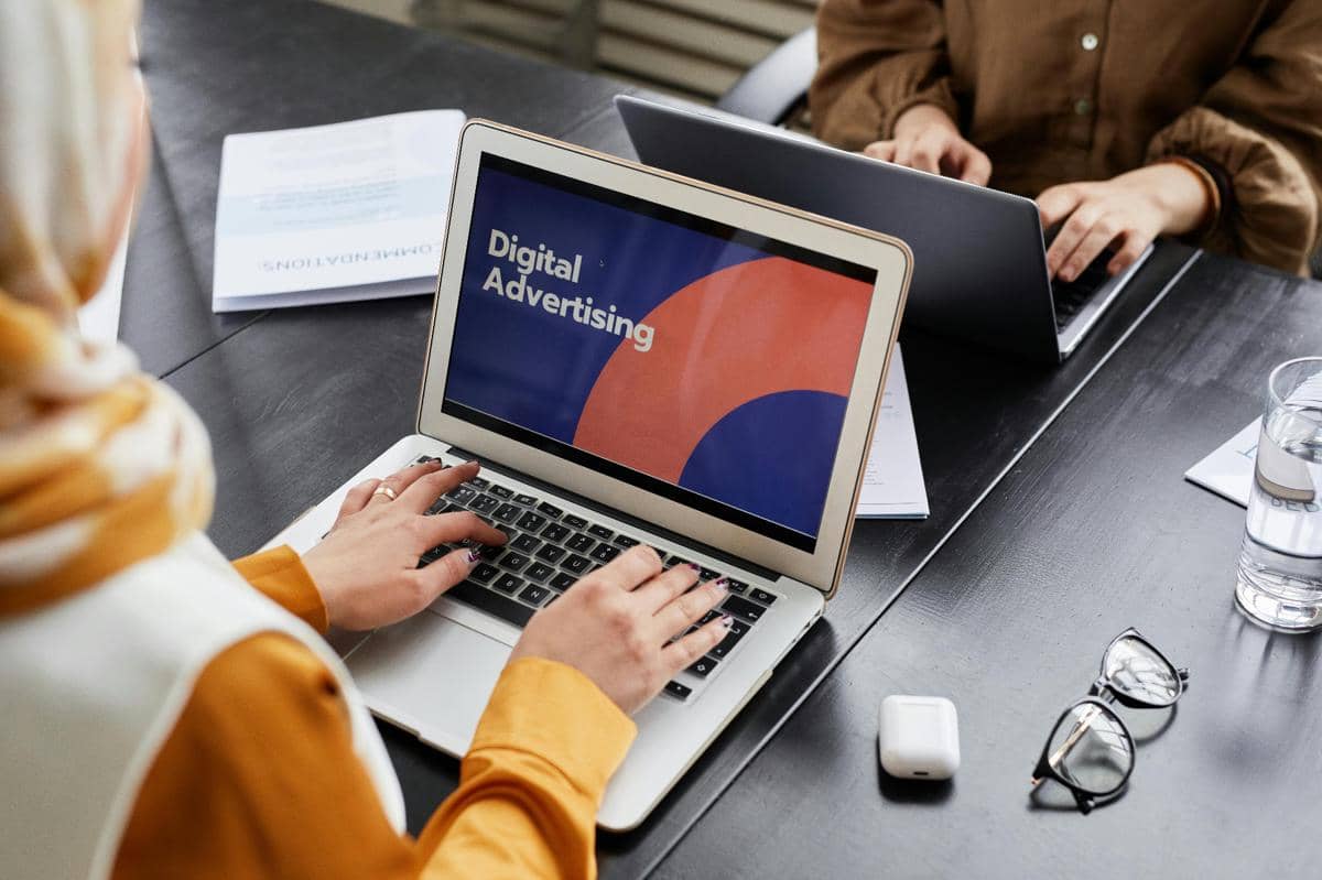 Seorang perempuan bekerja di depan laptop dengan layar bertuliskan Digital Advertising di meja kerja bersama rekan lainnya.