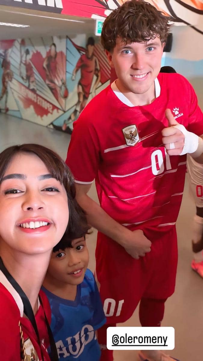 Seorang wanita dan anak kecil berfoto bersama pemain sepak bola berseragam merah di area stadion dengan mural bertema olahraga.