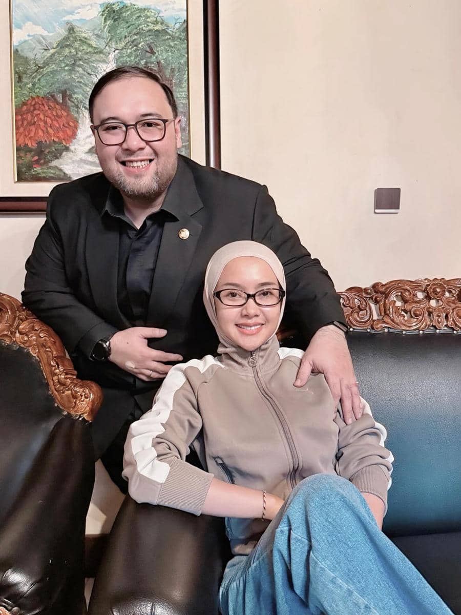 Seorang pria mengenakan jas hitam tersenyum berdiri di belakang seorang wanita berhijab dan berkacamata yang duduk di sofa kulit hitam.