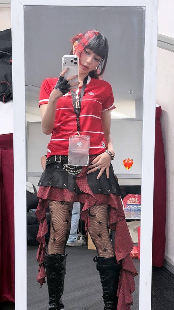 Seorang perempuan berambut pendek dengan highlight merah berpose di depan cermin mengenakan jersey merah, rok asimetris, dan sepatu bot hitam.
