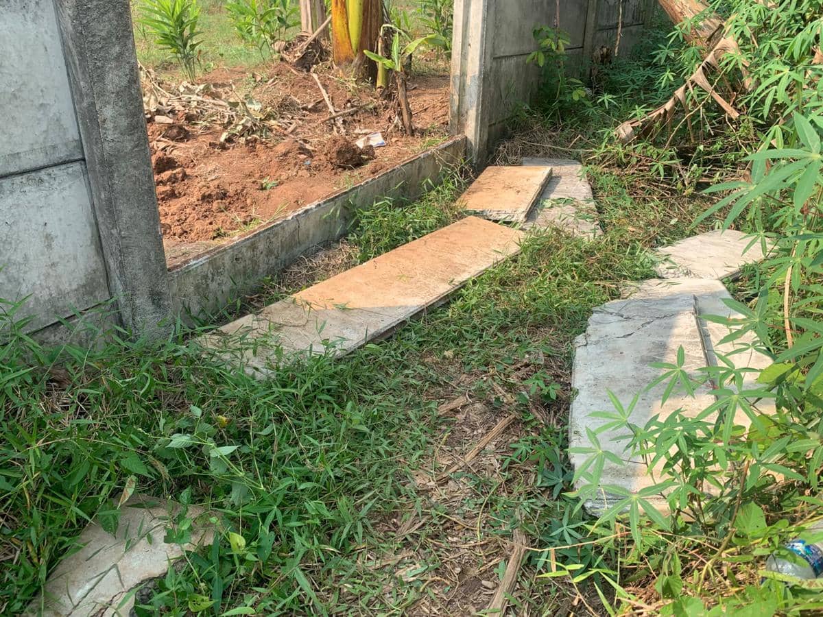 Tembok beton di area perumahan dengan bagian yang jebol dan pecahan semen berserakan di tanah berumput.