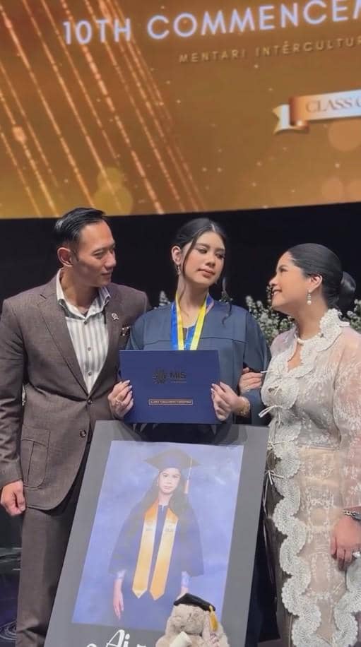 Seorang siswi mengenakan toga wisuda memegang ijazah didampingi dua orang dewasa di acara wisuda sekolah menengah.