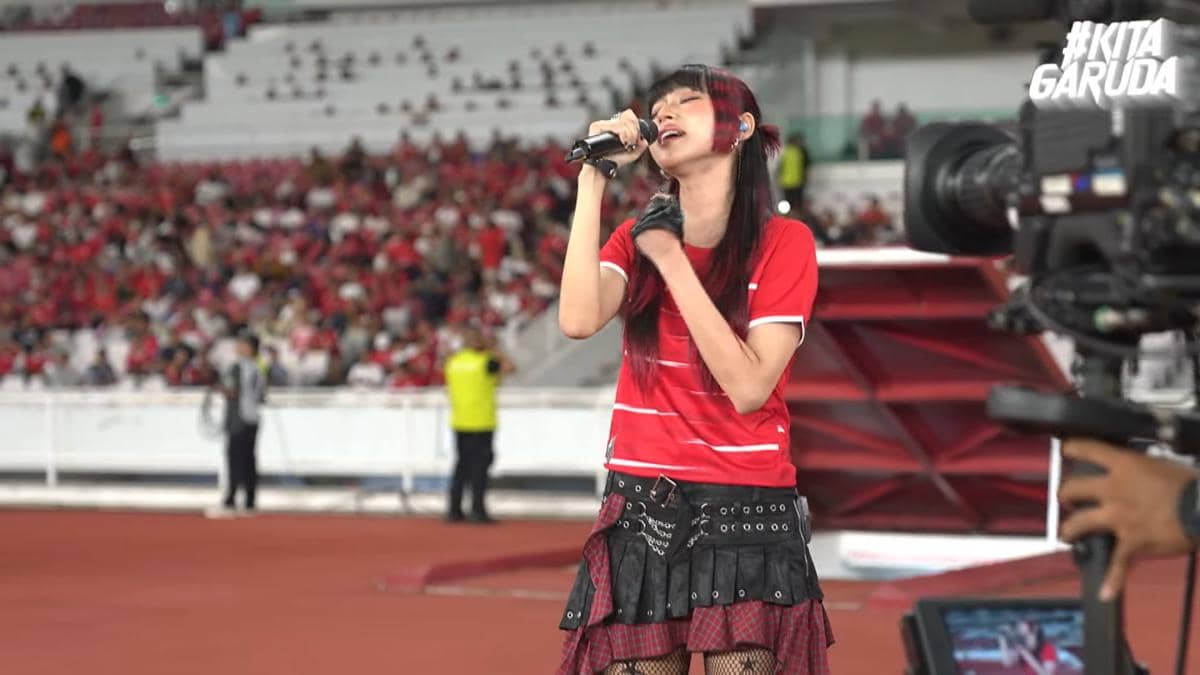 Seorang penyanyi mengenakan kostum merah tampil di stadion sambil memegang mikrofon di depan penonton sebelum pertandingan sepak bola.