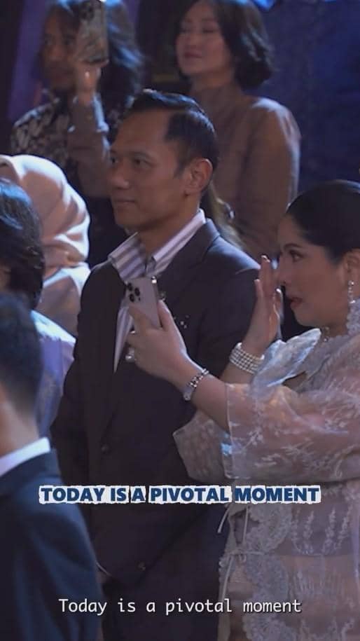 Seorang pria dan wanita berpakaian formal menghadiri acara wisuda, wanita tampak terharu sambil memegang ponsel untuk merekam momen.