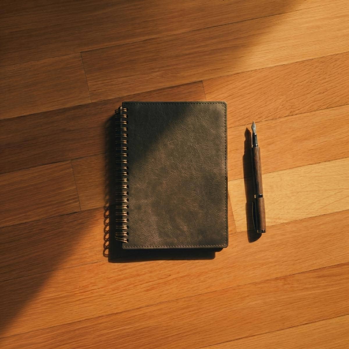 Sebuah buku catatan kulit berwarna gelap dengan spiral emas dan pena kayu diletakkan di lantai kayu yang diterangi cahaya hangat.
