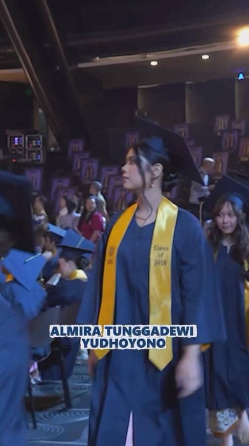 Seorang siswi mengenakan toga wisuda berwarna biru tua dengan selempang kuning berjalan di acara kelulusan SMA di dalam aula.