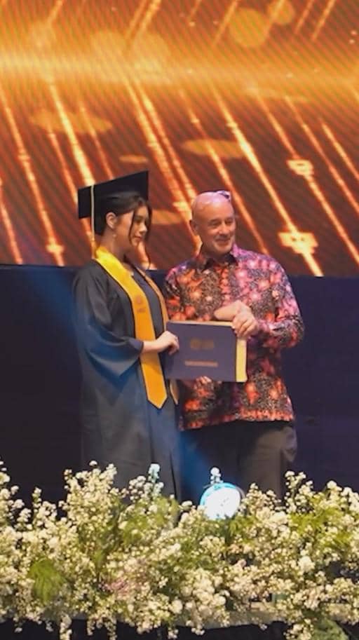 Seorang siswi mengenakan toga wisuda menerima ijazah dari seorang pria di atas panggung dengan latar pencahayaan emas.