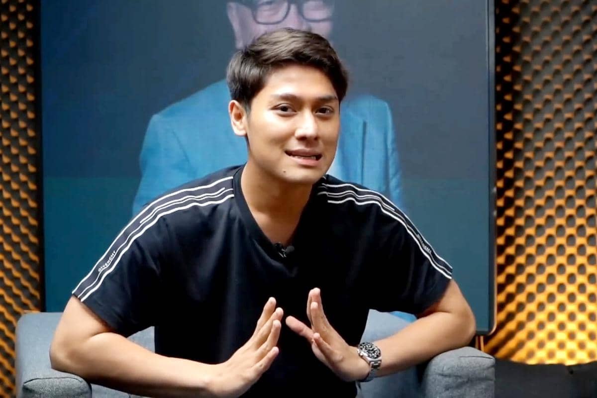 Rizky Billar mengenakan kaus hitam bergaris putih sedang berbicara di studio dengan latar belakang dinding bertekstur dan layar besar.