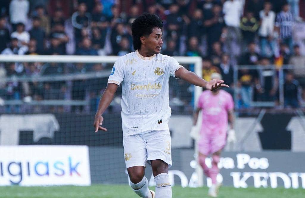 Seorang pemain Arema FC mengenakan seragam putih sedang berada di lapangan dengan latar belakang penonton di stadion.
