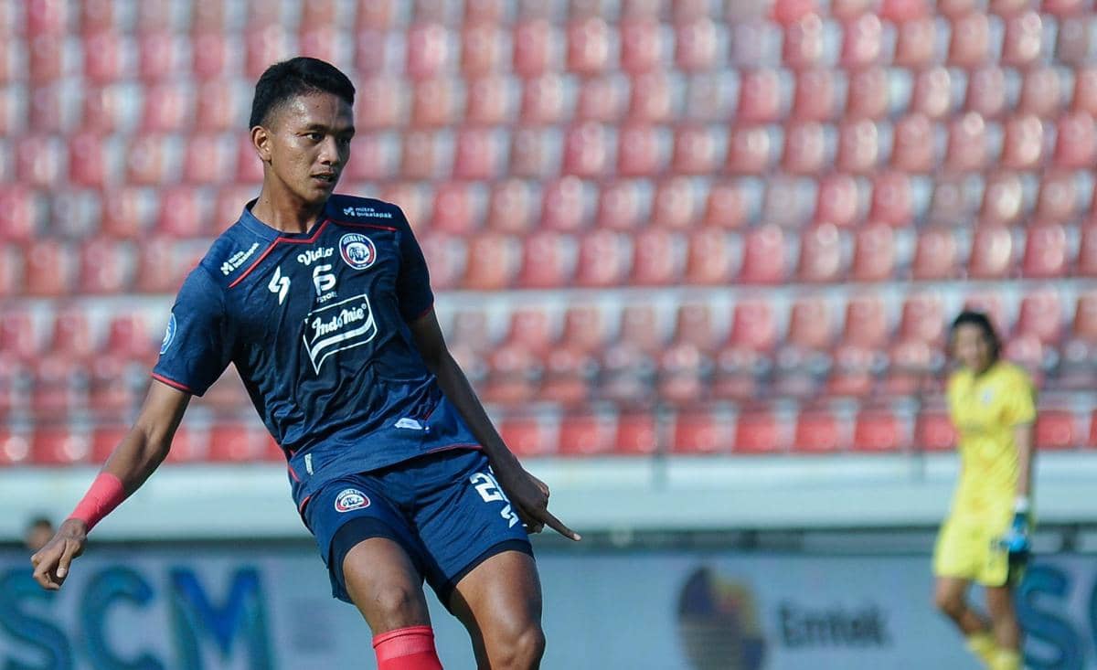 Seorang pemain Arema FC mengenakan seragam biru tengah mengontrol bola di lapangan dengan latar tribun stadion kosong.