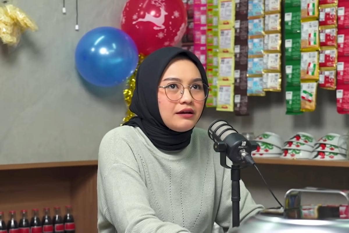 Salma Salsabil mengenakan kacamata dan sweater abu-abu sedang berbicara di depan mikrofon di sebuah studio podcast.