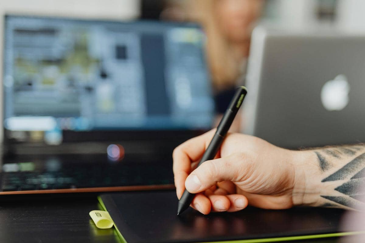 Seorang desainer menggunakan pena digital di tablet grafis dengan laptop terbuka di meja kerja untuk membuat desain.