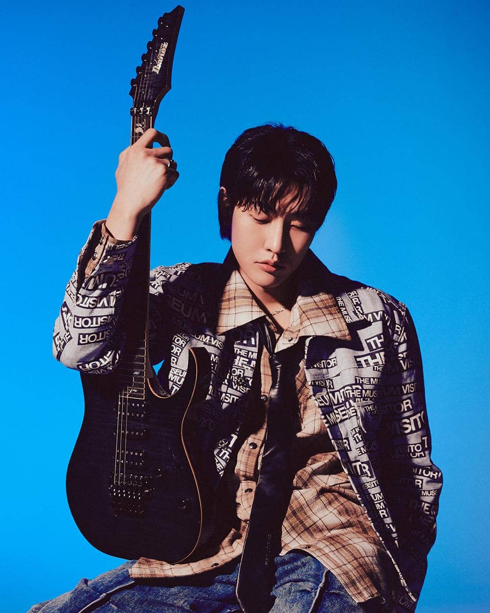 Seorang pria berpose sambil memegang gitar listrik hitam di depan latar belakang biru dengan pencahayaan artistik.