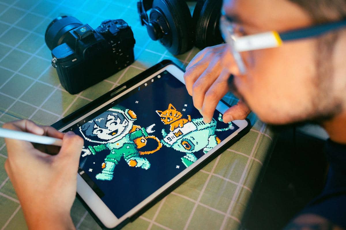 Seorang ilustrator sedang menggambar karakter bergaya pixel art di tablet digital dengan stylus di atas meja kerja.