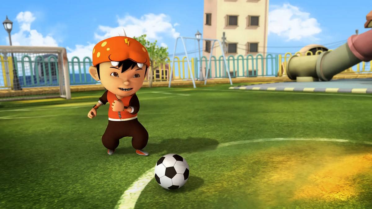 Karakter animasi Boboiboy mengenakan topi oranye sedang bermain sepak bola di lapangan hijau dengan latar taman bermain.