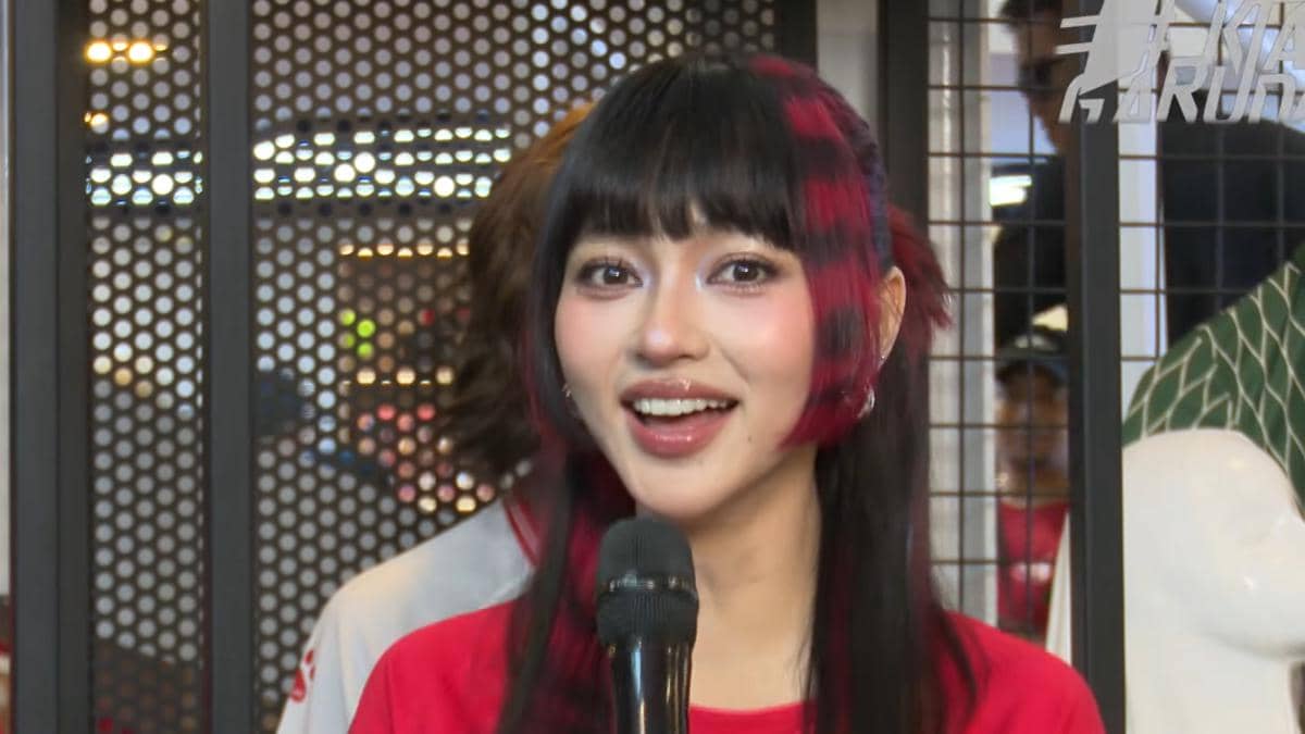 Seorang perempuan berambut hitam dengan highlight merah memegang mikrofon dan tersenyum di area wawancara menjelang pertandingan timnas.