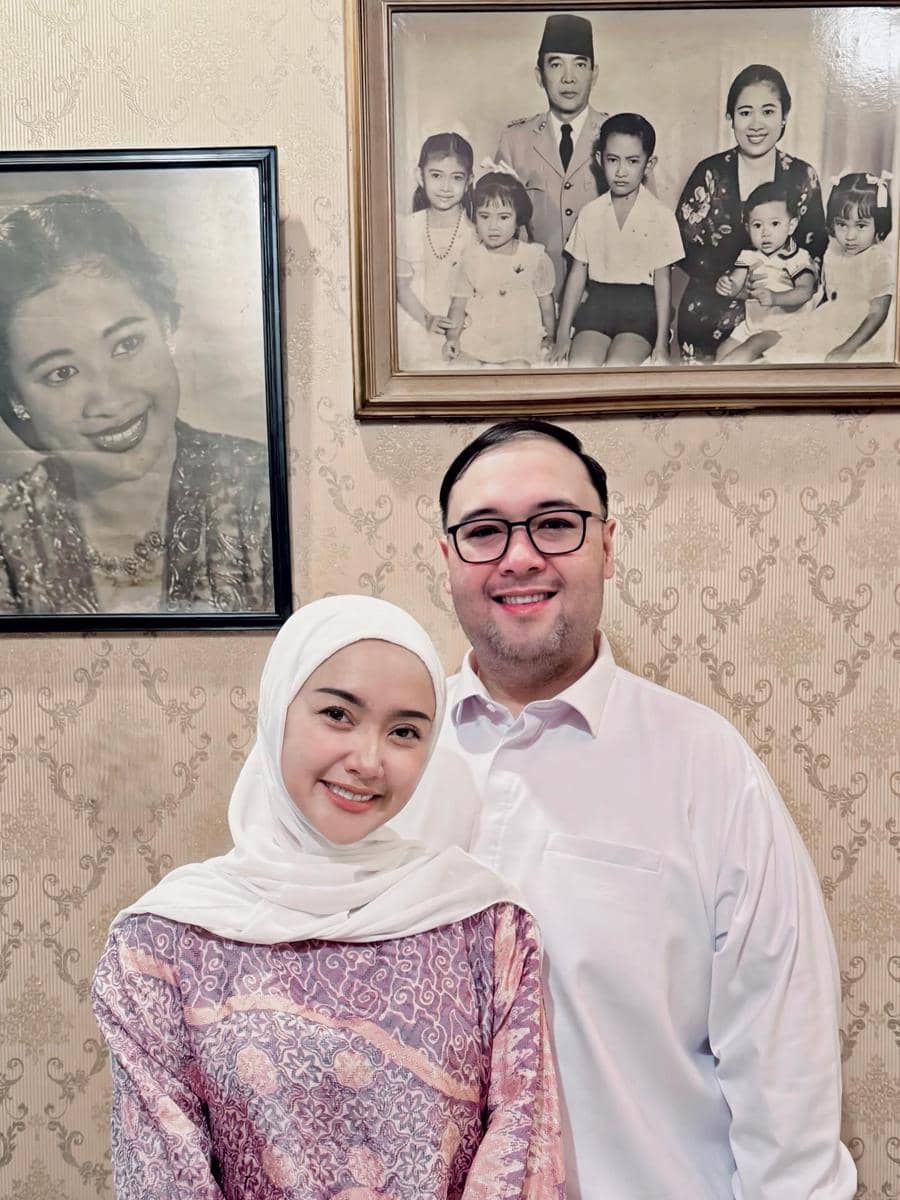 Seorang perempuan berhijab dan seorang pria berkacamata berpose bersama di depan dinding dengan foto keluarga hitam putih.