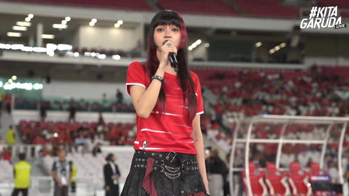 Seorang penyanyi mengenakan kaus merah sedang tampil di stadion dengan mikrofon di tangan menjelang pertandingan sepak bola.