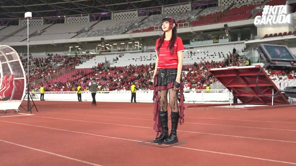 Seorang perempuan mengenakan kostum merah dan hitam berdiri di tepi lapangan stadion dengan penonton di tribun belakangnya.